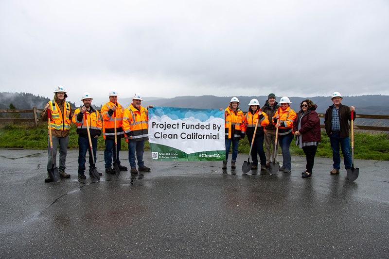 Caltrans staff and local dignitaries stand on each sign of a banner which reads Project Funded by Clean California. They stand on a paved parking lot at Berry Summit in Humboldt County with a mountain ridge behind them in the distance.