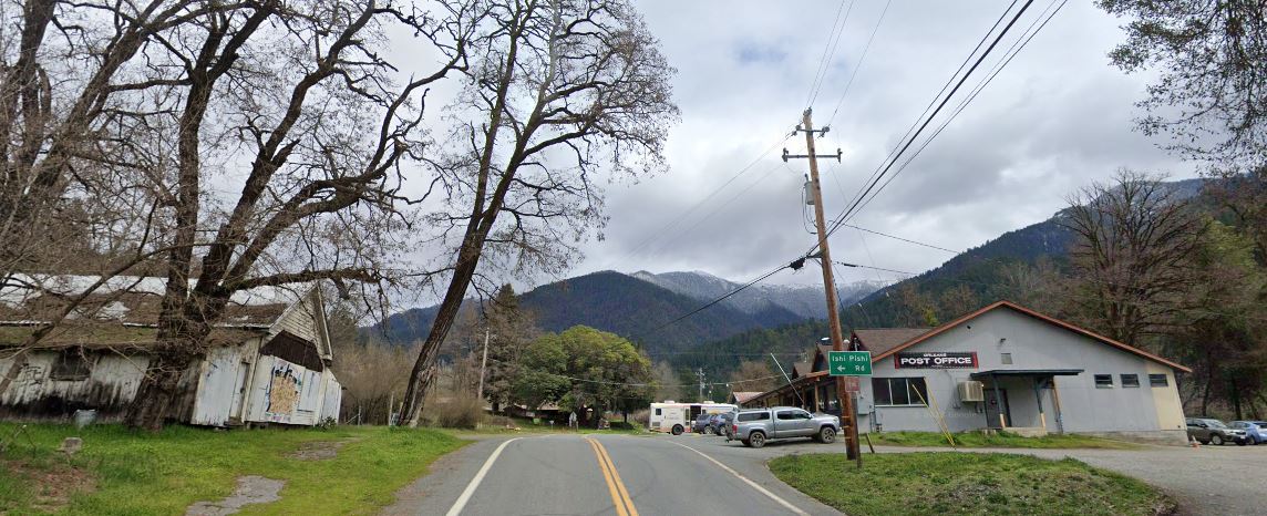 State route 96 extends from the foreground to the background with tree covered mountains in the distance. A grey-blue cloud bank sits over the mountains. On one side of the road, sits a dilapidated building surrounded by trees. On the other side is a small building with a sign that reads Orleans post office. A road sign indicating Ishi Pishi Road stands near the Post Office. 