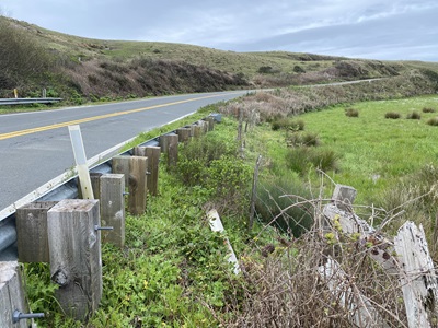 Route 1 near Point Arena California in Mendocino County. A two-lane highway is flanked by grass covered hills. It extends from the left to right with a grey cloudy sky.