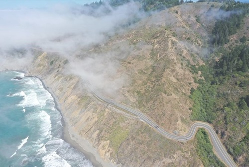 Aerial photo of Route 1 near Westport in Mendocino County. The road winds along the coast with the ocean in the foreground. Fluffy white clouds obscure part of the photo. 