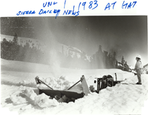 A black and white picture of a snowblower driving through snow that's nearly as tall as the vehicle. A Caltrans employee stands to the side of the vehicle on top of the snow. Writing on the image reads "June 1 , 1983 at Gate. Sierra Daily News."