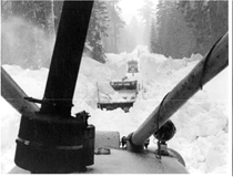 A black and white image from the point of view of a snowplow showing a snow blower removing snow from State Route 120 W to Yosemite in 1983. A man walks toward another vehicle in the distance.