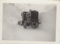 An undated black and white image of a truck with snow chains on next to a wall of snow.