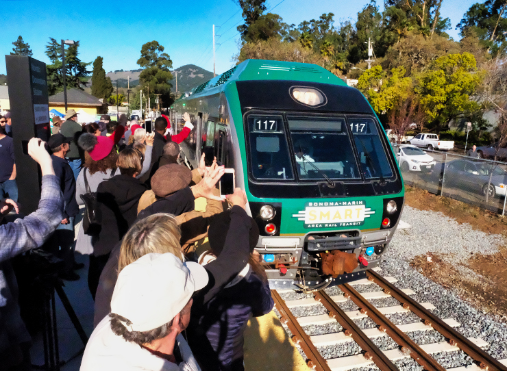 Photo of the SMART train pulling into the Novato station on opening day. A crowd of cheering people is in the foreground.